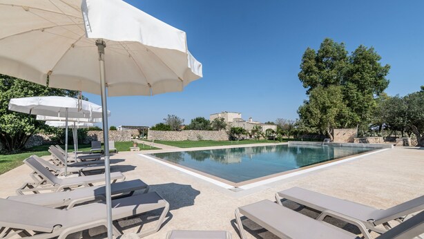 Area piscina con sedie a sdraio e ombrelloni, una piscina limpida e una vista su un edificio storico sullo sfondo.