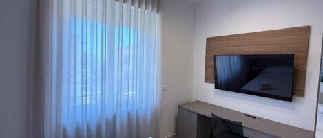 Une chambre d’hôtel moderne équipée d’un lit, d’un bureau, d’une chaise, d’un téléviseur à écran plat fixé au mur et d’une grande fenêtre avec des rideaux légers.