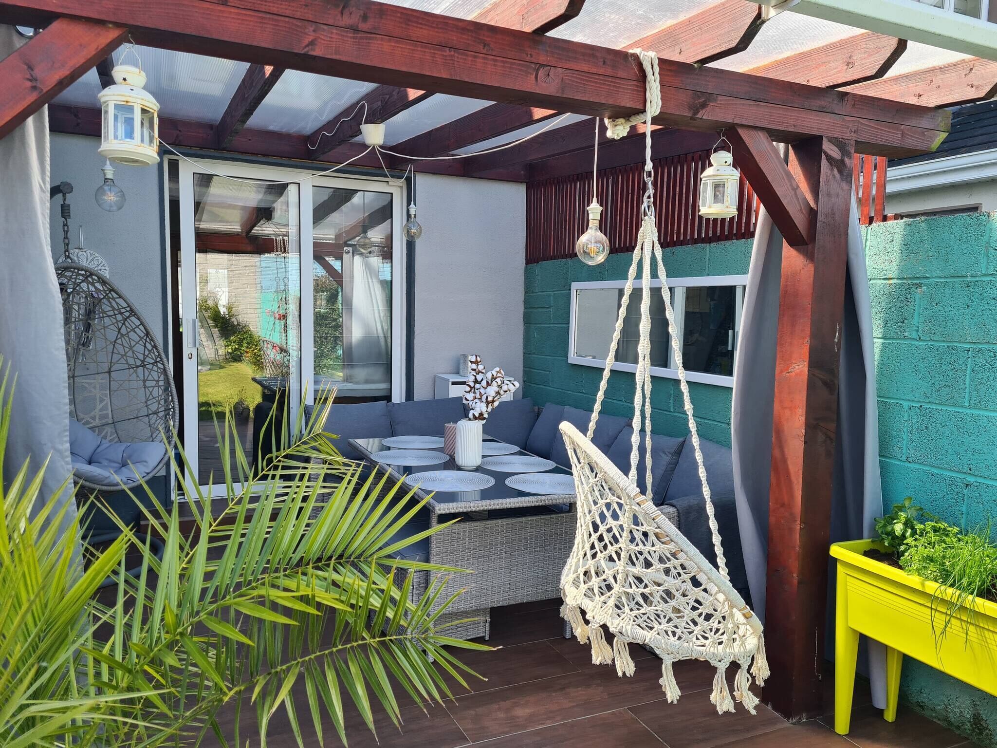 A cozy patio area with a cushioned seating arrangement, a hanging macrame chair, and a yellow planter with greenery.