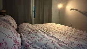 Quarto com cama coberta por roupa de cama floral, janela com cortinas e luminária na parede.