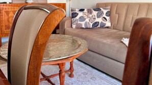 Uma área de refeições com uma cadeira de madeira, uma mesa redonda, um sofá com almofadas estampadas e um quadro na parede.