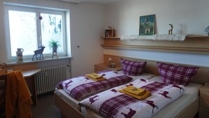 A bedroom with two beds, a desk, a chair, a radiator, a window with a view of trees, and a shelf with decorative items. - AllgäuZeit apartment with indoor pool & sauna & free WLAN (Missen-Wilhams)
