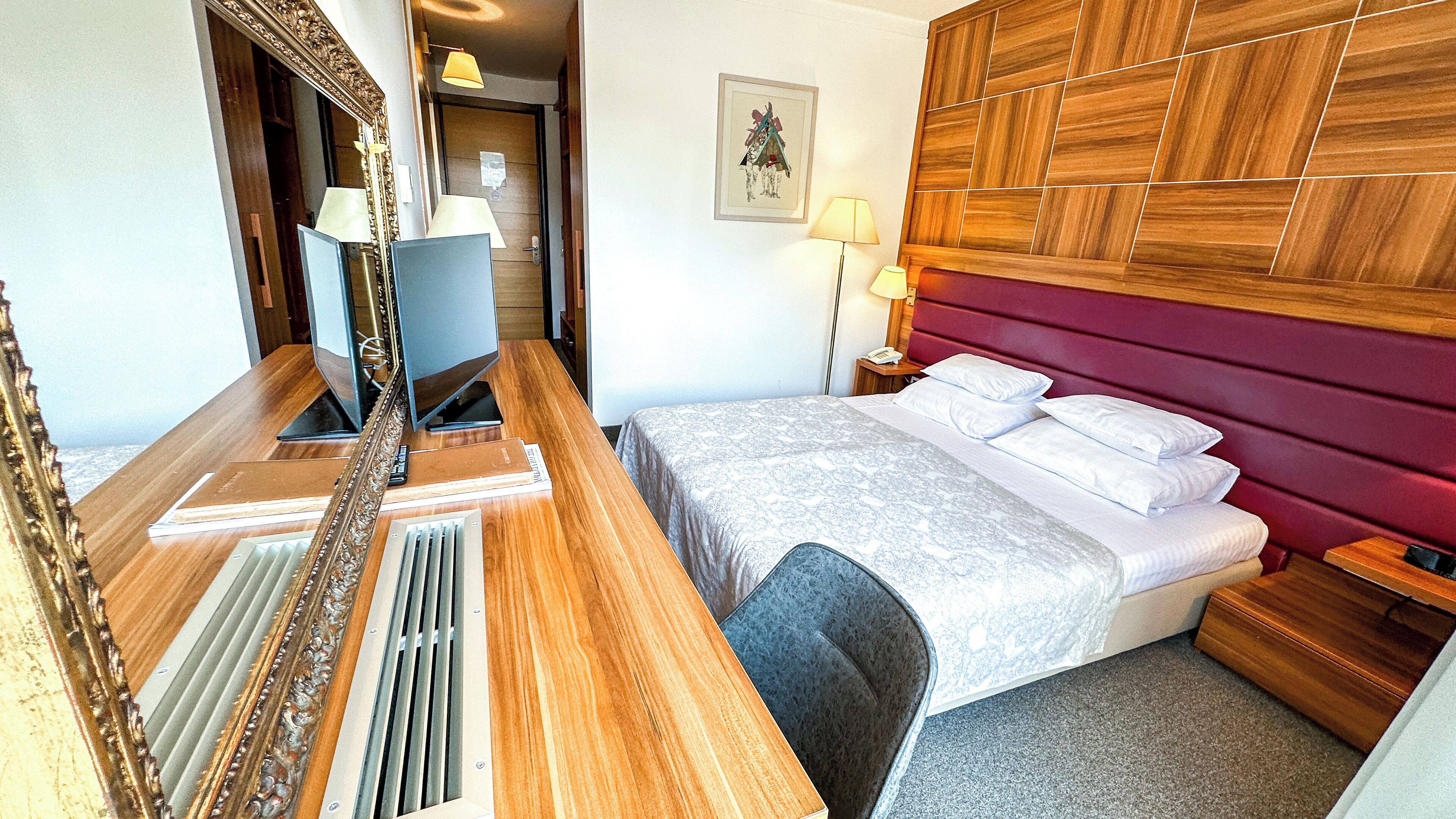 A hotel room with a wooden desk, a bed with a red headboard, a framed picture on the wall, and a lamp on the nightstand.