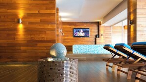 Un espacio de spa moderno con suelo de madera, un elemento circular con mosaicos, un televisor en la pared y sillones reclinables acolchados de color azul.
