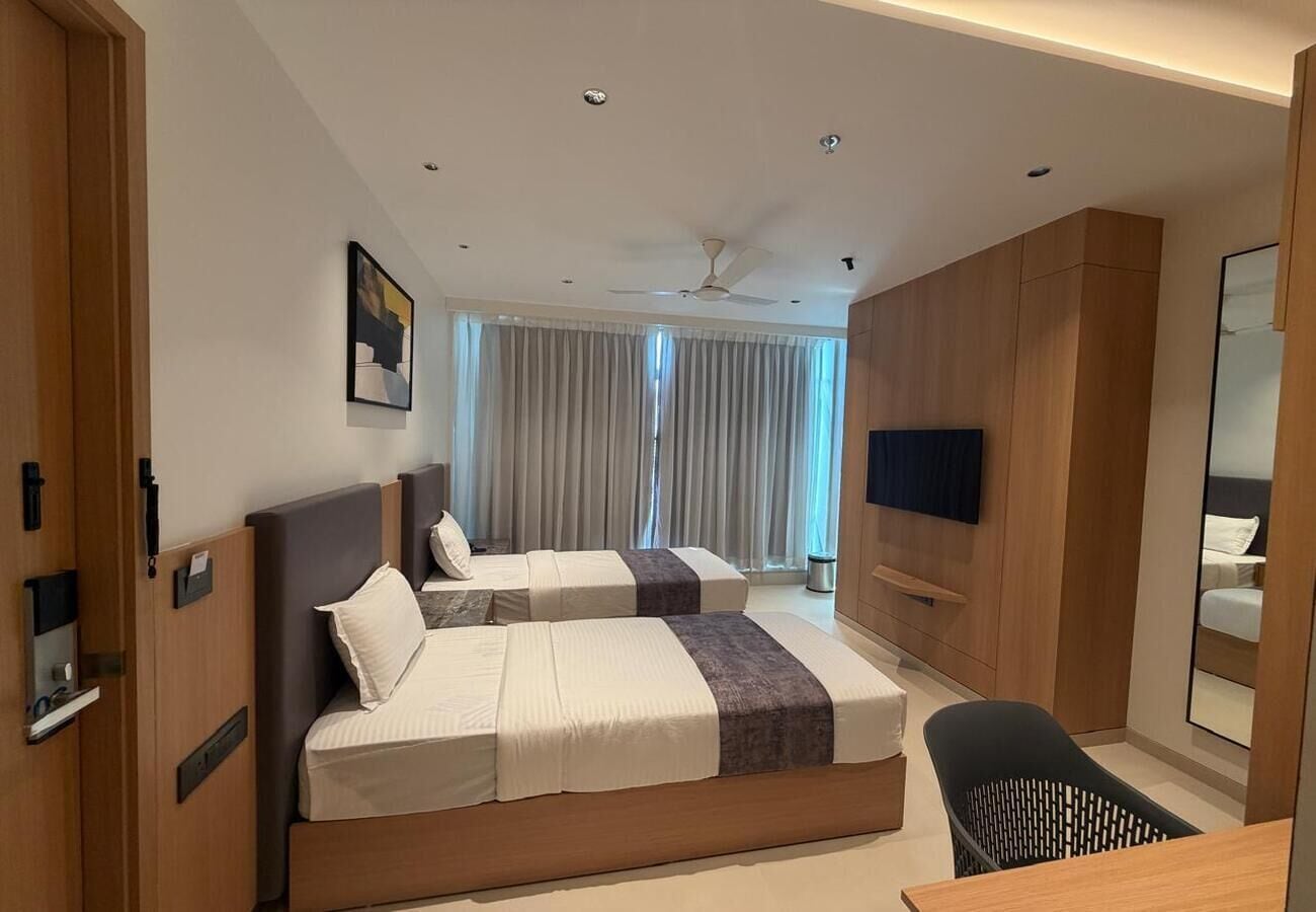 A hotel room with two beds, a desk, a chair, a TV, and a ceiling fan.