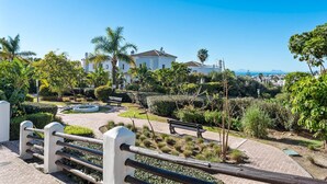 A landscaped garden with a pathway, benches, and a white building with a tiled roof. - Perla Del Sol Estepona (Estepona)