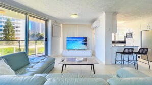 Ein modernes Wohnzimmer mit großem Fernseher, einer Couch, einem Couchtisch und einer Küchenzeile inklusive Mikrowelle und Kühlschrank.
