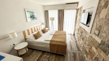 A hotel room with a bed, a small round table, a lamp, a TV, and a stone wall.