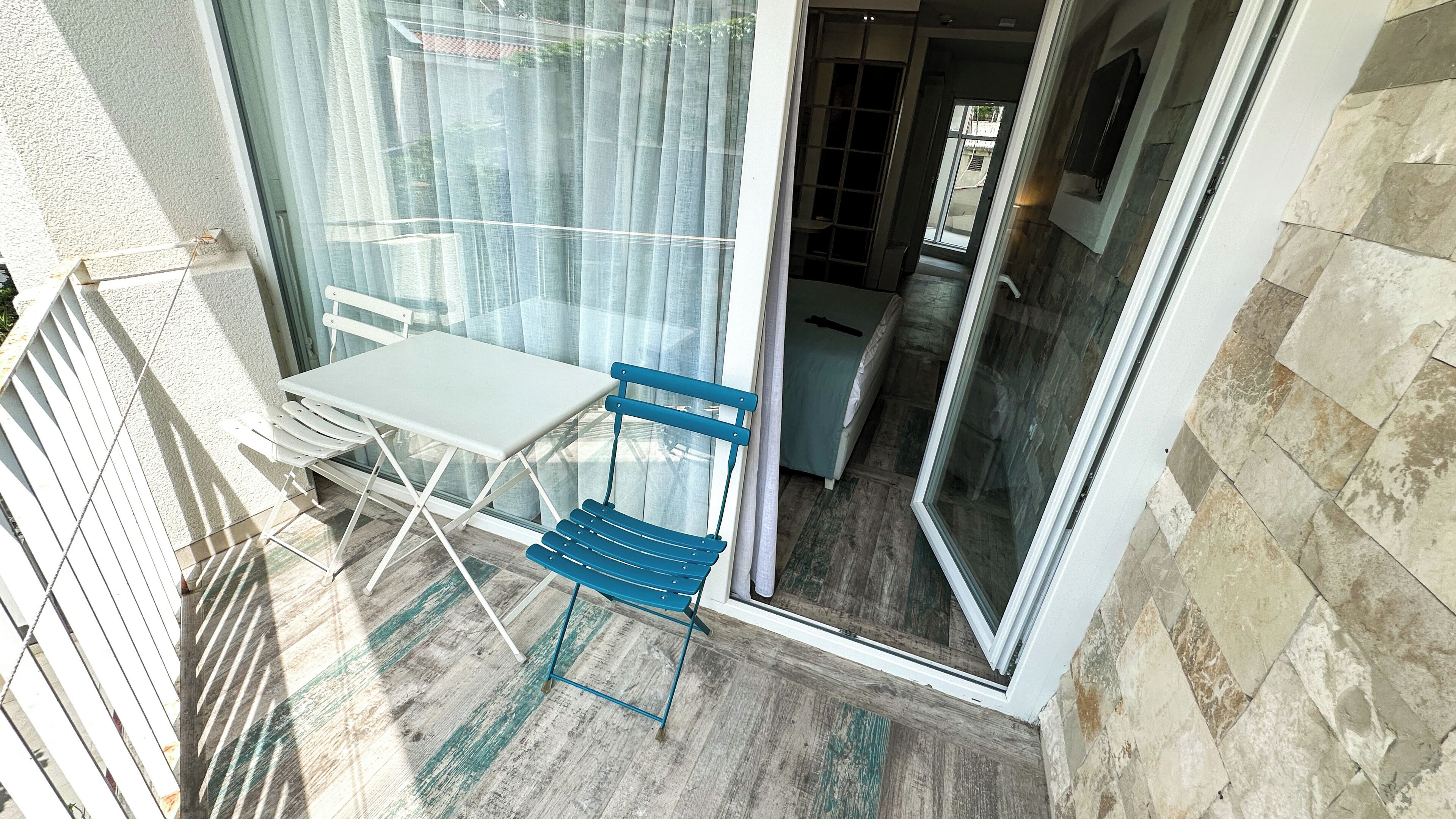A balcony with a white table and blue chairs, a glass door, and a stone wall.