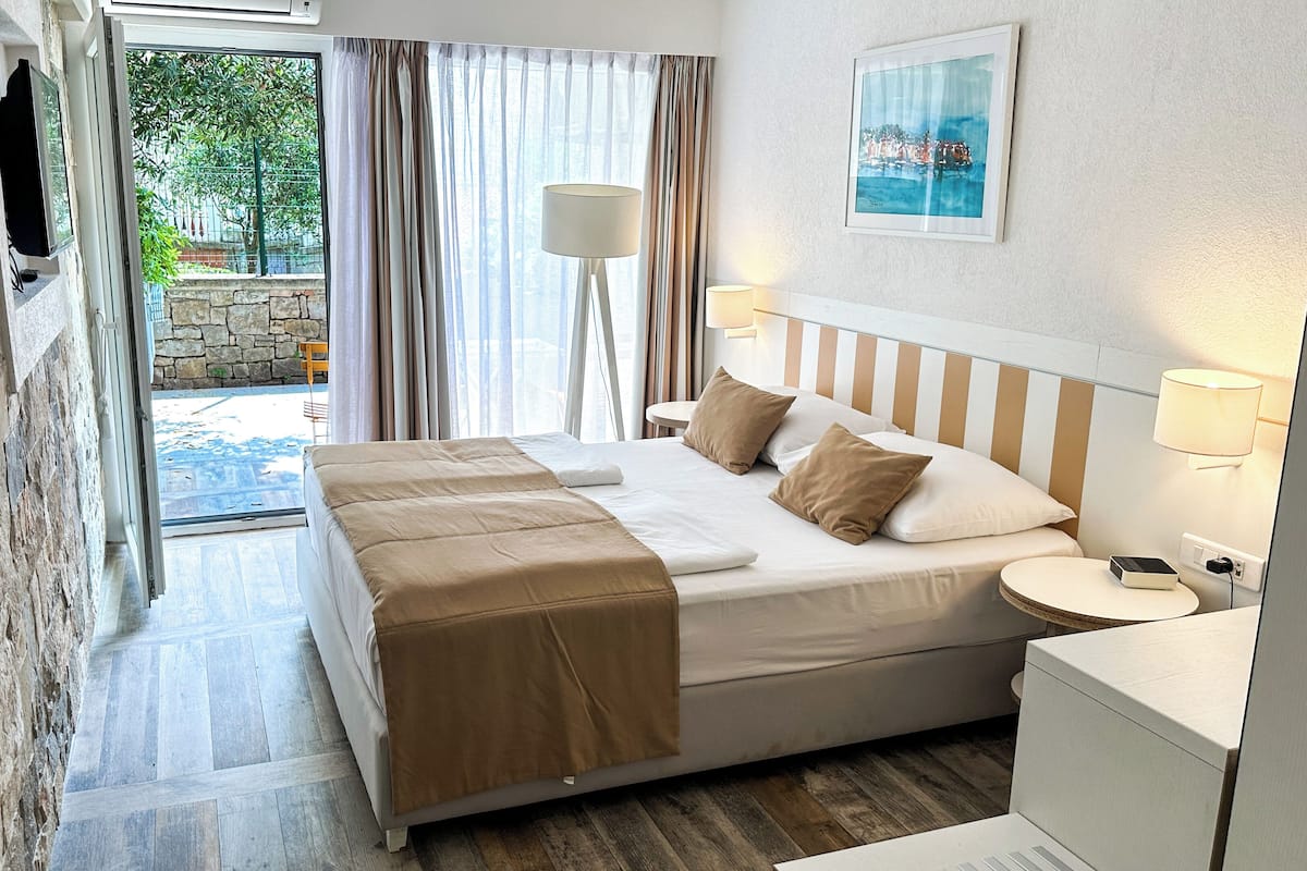 Ein Hotelzimmer mit Bett, Nachttischen, einer Lampe, einem Gemälde und Blick ins Freie.