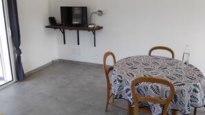 Een kleine kamer met een tafel en stoelen, een televisie op een plank en een raam met gordijnen.