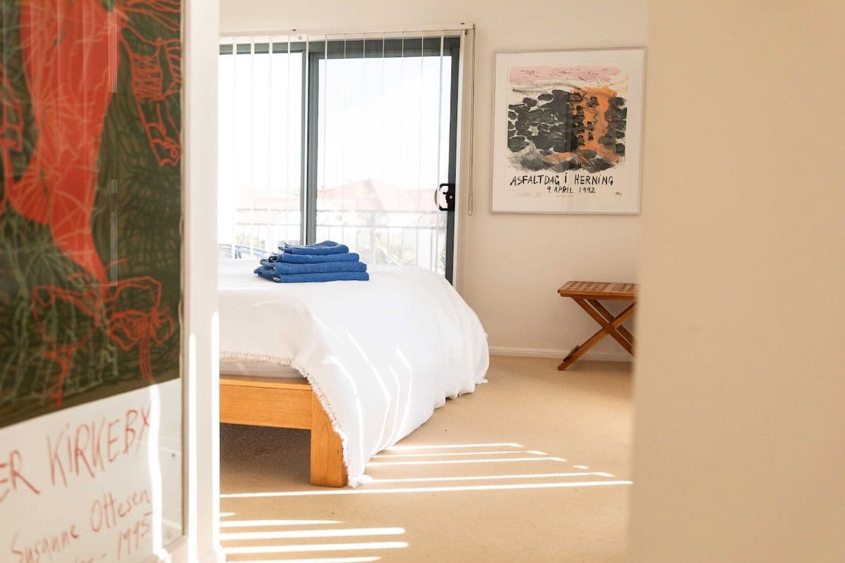 Un dormitorio con una cama, una ventana, una obra de arte enmarcada y un banco de madera.