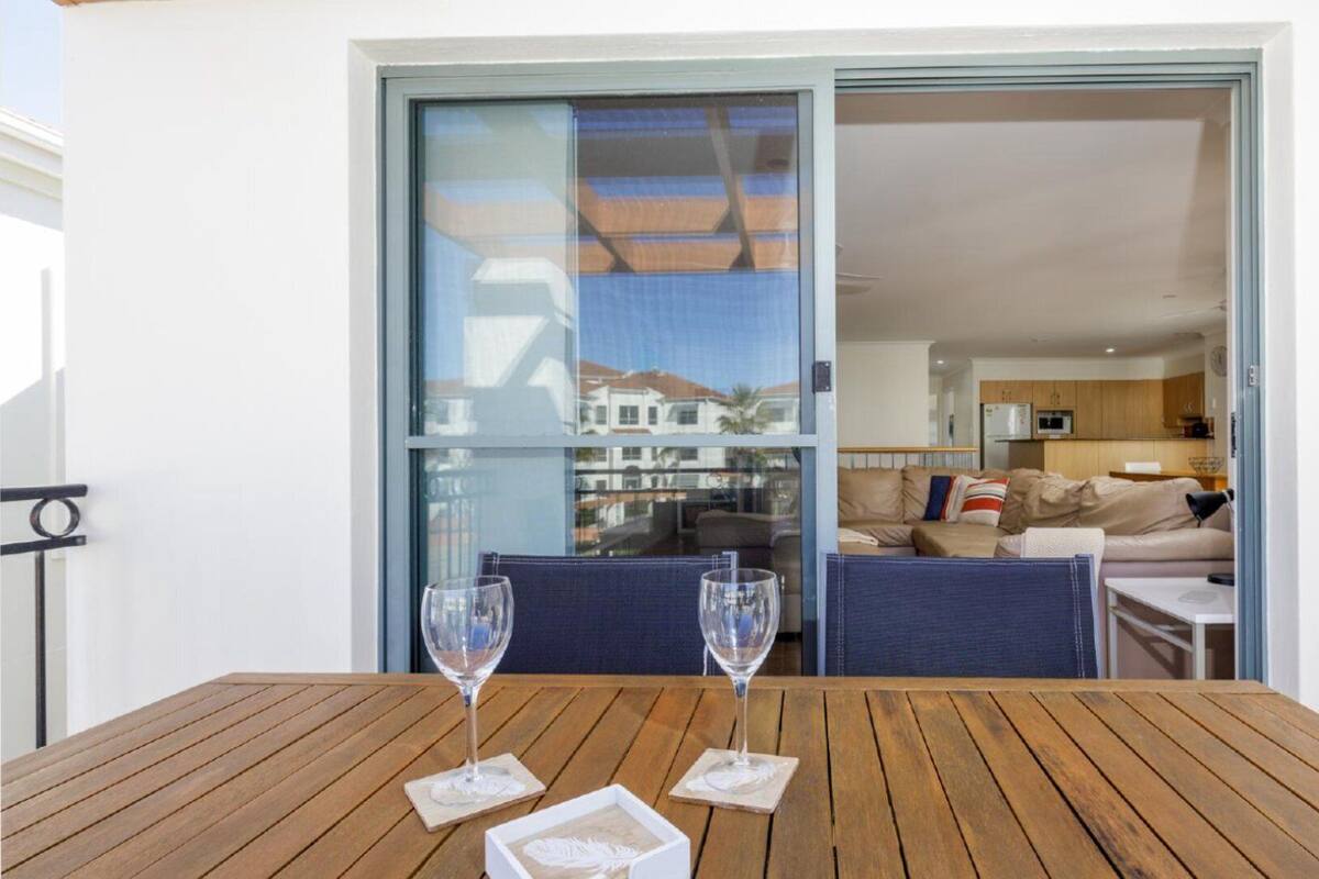 Una mesa de madera con dos vasos sobre posavasos, una puerta corredera de vidrio que da a una sala de estar con un sofá y una cocina visible a través de la puerta.