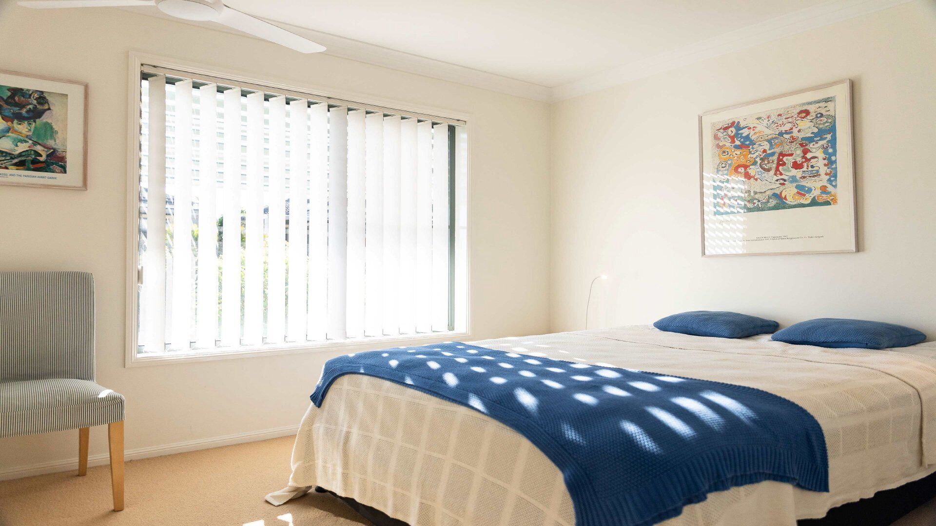 A bedroom with a bed, a chair, a window with blinds, and two wall-mounted artworks.
