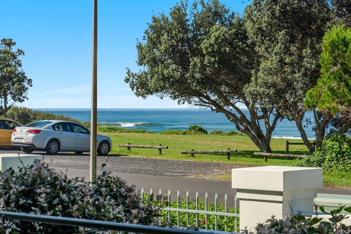 Vista costera con un coche blanco estacionado al borde de una carretera, una valla de madera y un árbol grande al frente.