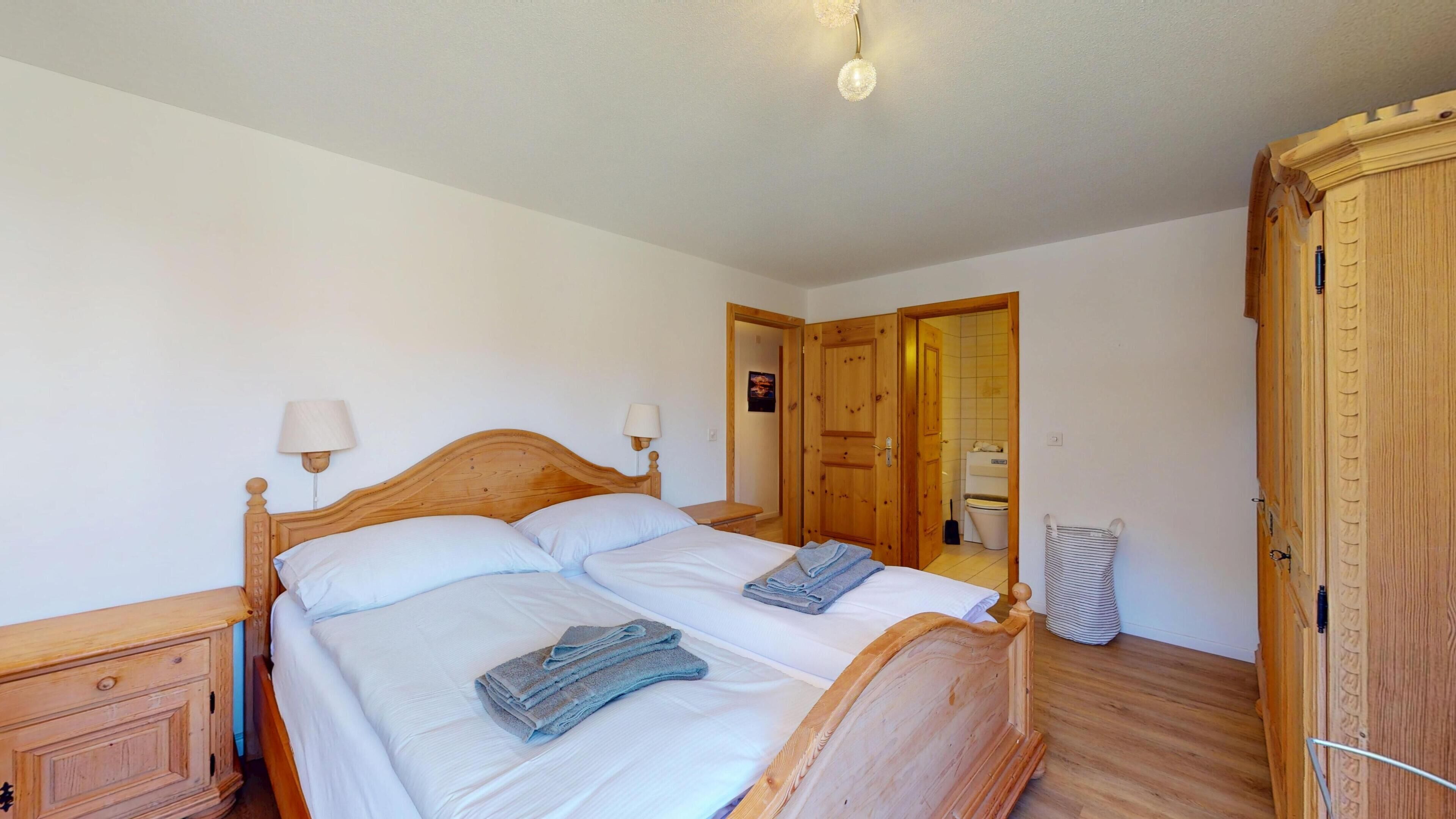 Un dormitorio con una cama de madera, dos mesitas de noche, un armario de madera y un baño visible a través de una puerta abierta.