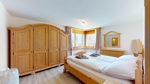 Une chambre avec une grande armoire en bois, un lit avec du linge de maison blanc, une fenêtre avec des rideaux et un miroir sur le mur.
