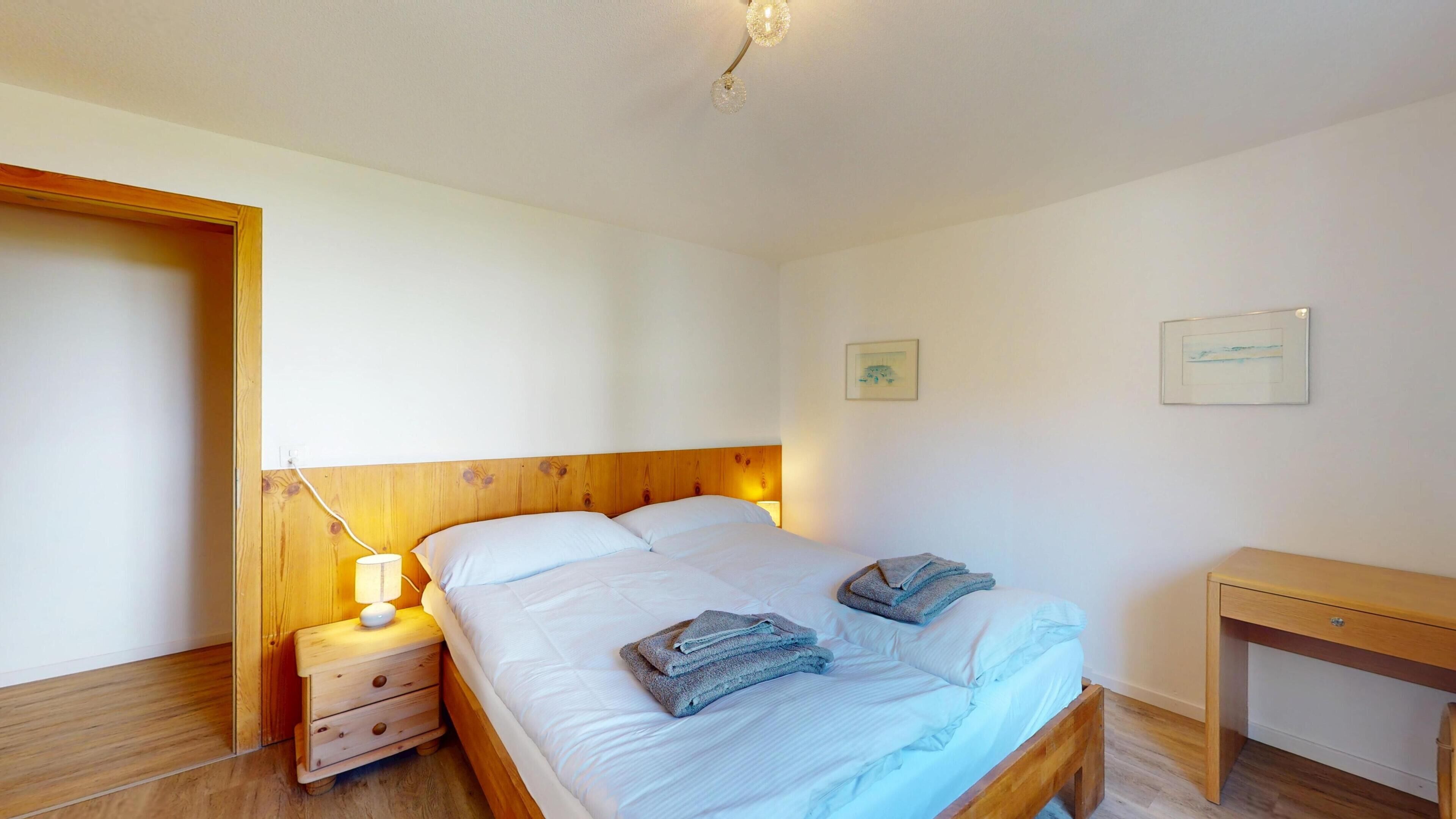 Quarto com cabeceira de madeira, mesa de cabeceira com abajur, cama com roupa de cama branca e duas toalhas dobradas.