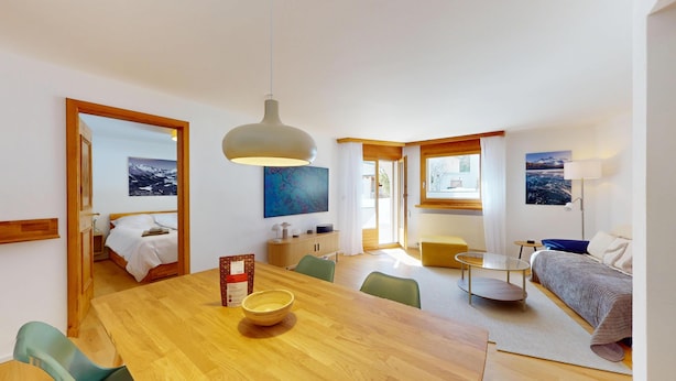 Una habitación de hotel moderna con una mesa de comedor de madera, un sofá, una pequeña mesa redonda y una cama con vistas al paisaje nevado.
