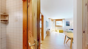 Un interior moderno con una puerta de madera, paredes con azulejos, una zona de comedor con mesa y sillas, y vistas por una ventana.