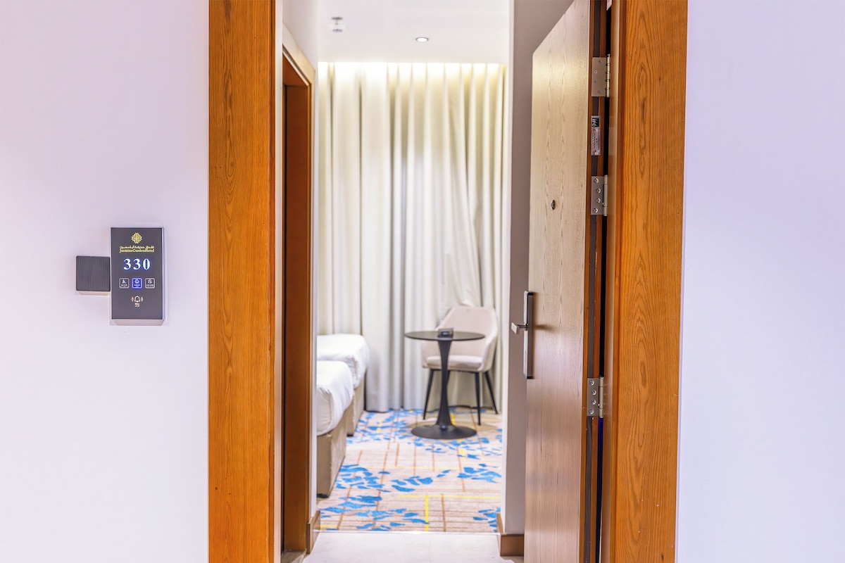 Een hotelkamer met een bed, een klein tafeltje en een stoel. Een bord op de muur geeft kamer nummer 330 aan.