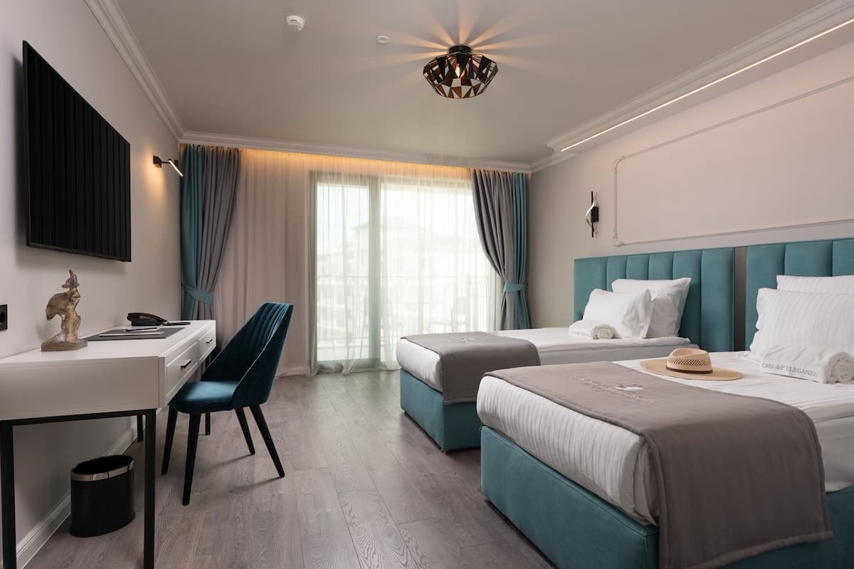 A hotel room with a desk, two beds, a chandelier, and a view of the outdoors.