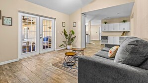 A spacious living room with a gray sofa, a wooden coffee table, and a patterned rug. There is a sliding glass door leading to a balcony with a blue chair. A ceiling fan is visible, and the room has light-colored walls and wooden flooring. - Sunshine, Salt Air & Poolside Living Awaits (Seminole)