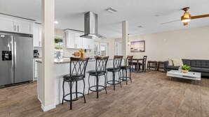 A modern kitchen with a stainless steel refrigerator, a central island with bar stools, and a dining area with a sofa and coffee table. - Sunshine, Salt Air & Poolside Living Awaits (Seminole)