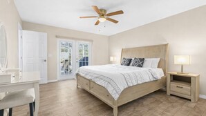 A bedroom with a wooden bed, a nightstand with a lamp, a ceiling fan, and a door leading to a balcony. - Sunshine, Salt Air & Poolside Living Awaits (Seminole)