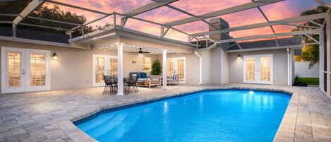 Una piscina con un porche techado, mobiliario exterior y vista al océano al atardecer.