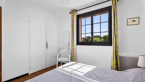 Un dormitorio con cama, una silla, una ventana con cortinas y una lámpara.