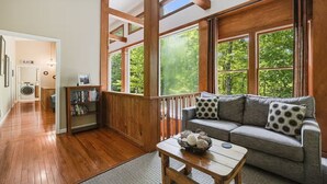 Ein gemütliches Wohnzimmer mit Holzbalken, einem grauen Sofa mit gemusterten Kissen, einem Holztisch und großen Fenstern mit Blick auf Grünflächen.