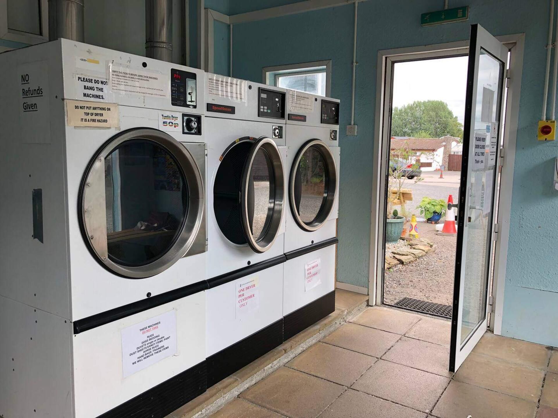 A laundry facility with three front-loading washing machines, a sign indicating 'NO Refunds Given,' and a door to the outside.