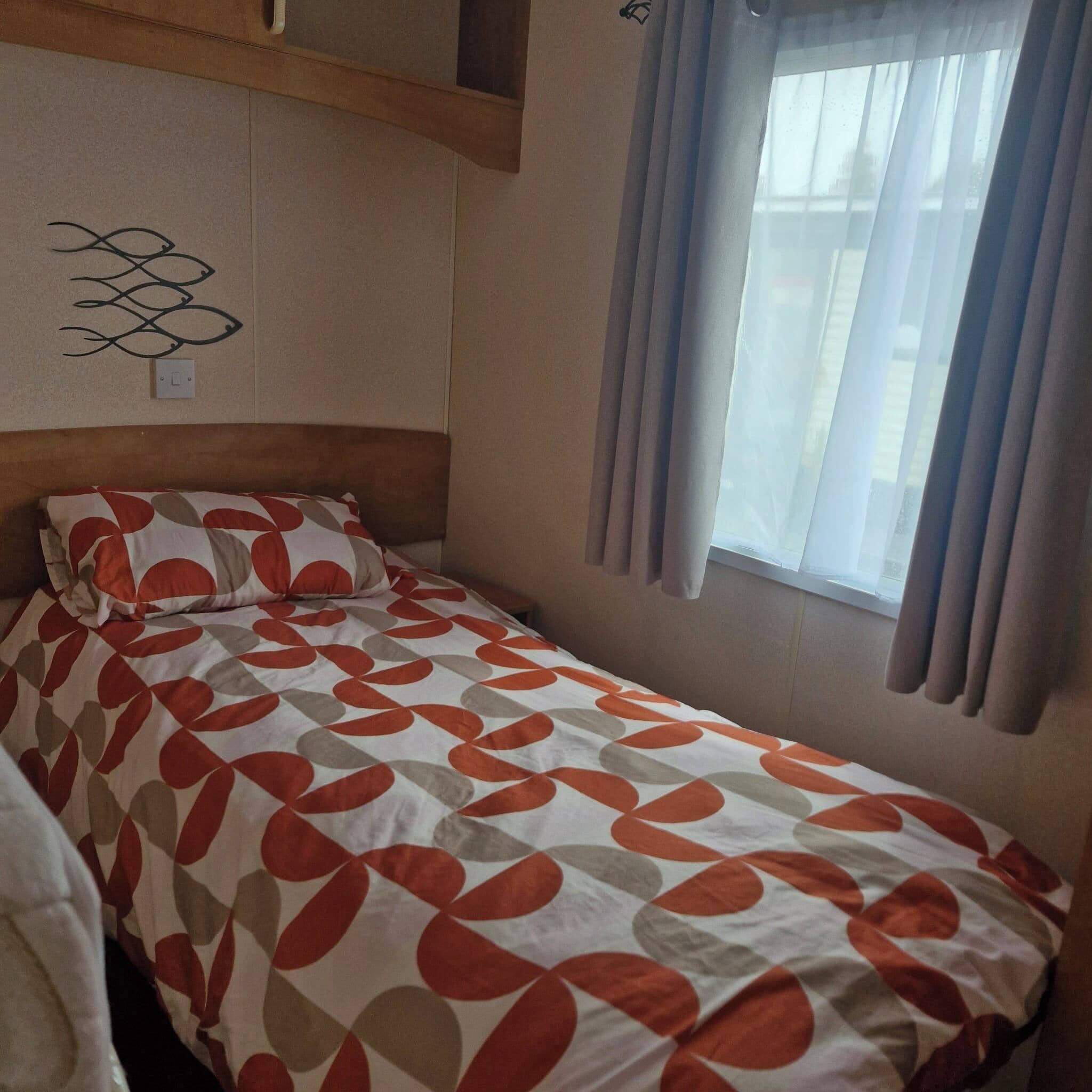 Un letto singolo con copriletto decorato, una testiera, un interruttore della luce fissato al muro e una finestra con tende.