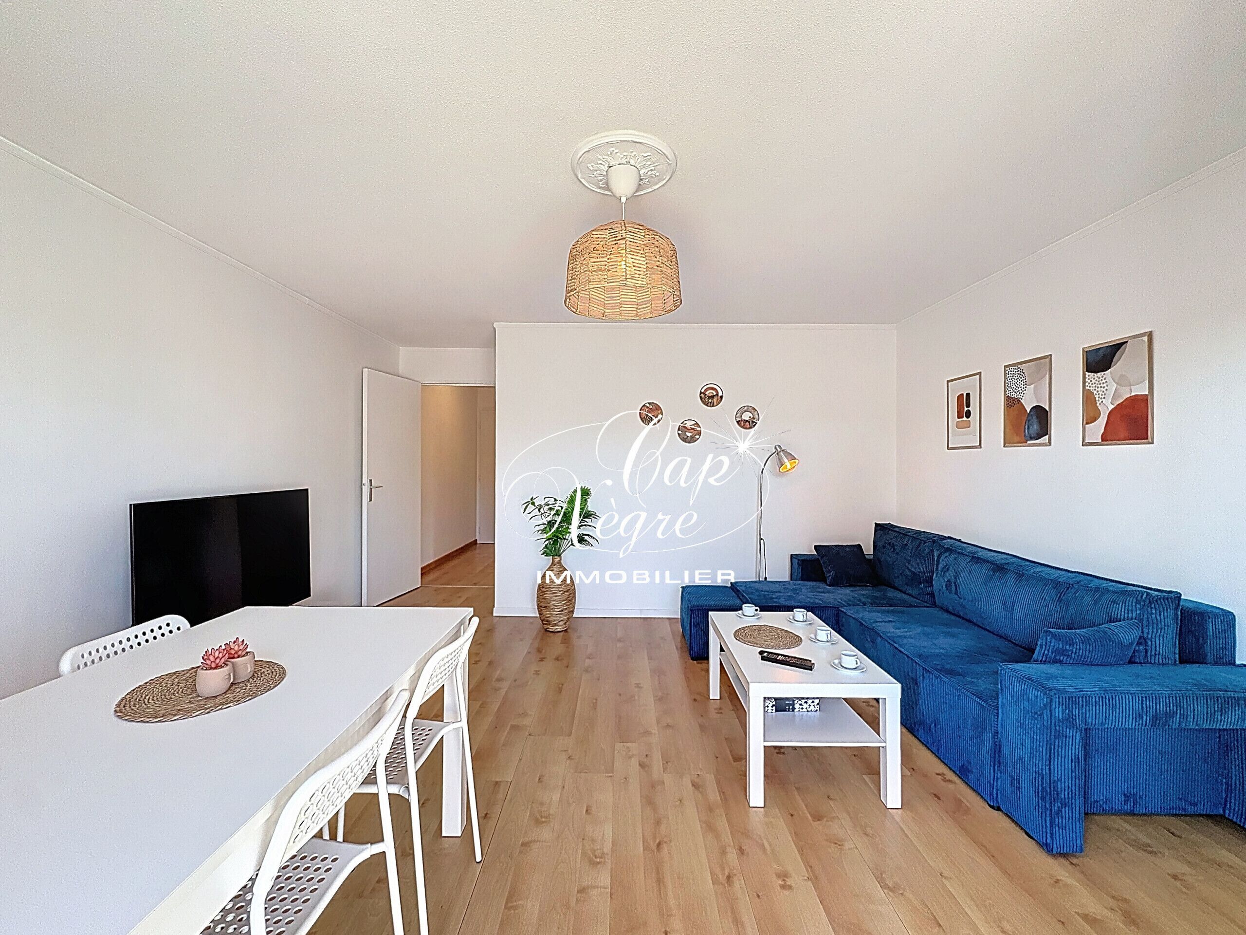 Un salon moderne avec un canapé bleu, une table basse blanche, une table à manger avec des chaises, un téléviseur et un mur décoratif agrémenté d’une plante.