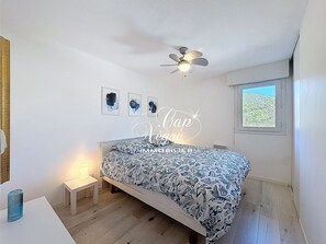 Ein Schlafzimmer mit Bett, Deckenventilator, Nachttisch, Fenster und Wandbildern.