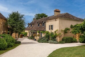 Um edifício de pedra tradicional com entrada por um caminho de cascalho, rodeado por sebes e árvores bem cuidadas.