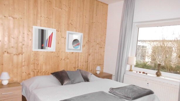 A bedroom with a wooden wall, a large bed with grey bedding, two bedside lamps, and a window with a view of trees. - At the meadow (Kuchelmiß)
