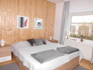 Un dormitorio con pared de madera, una cama grande con ropa de cama gris, dos lámparas de noche y una ventana con vistas a árboles.