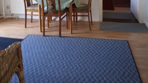A dining area with a table and chairs, a radiator, a painting on the wall, and a doorway leading to another room. - At the meadow (Kuchelmiß)