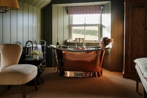 A room with a vintage copper bathtub, a plush armchair, a small table with a bottle and flowers, and a window with a view of greenery.