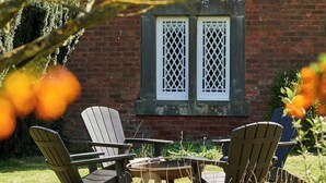 Een terras met Adirondack-stoelen en een tafel, een bakstenen gebouw met lattenramen, en een groen gazon.