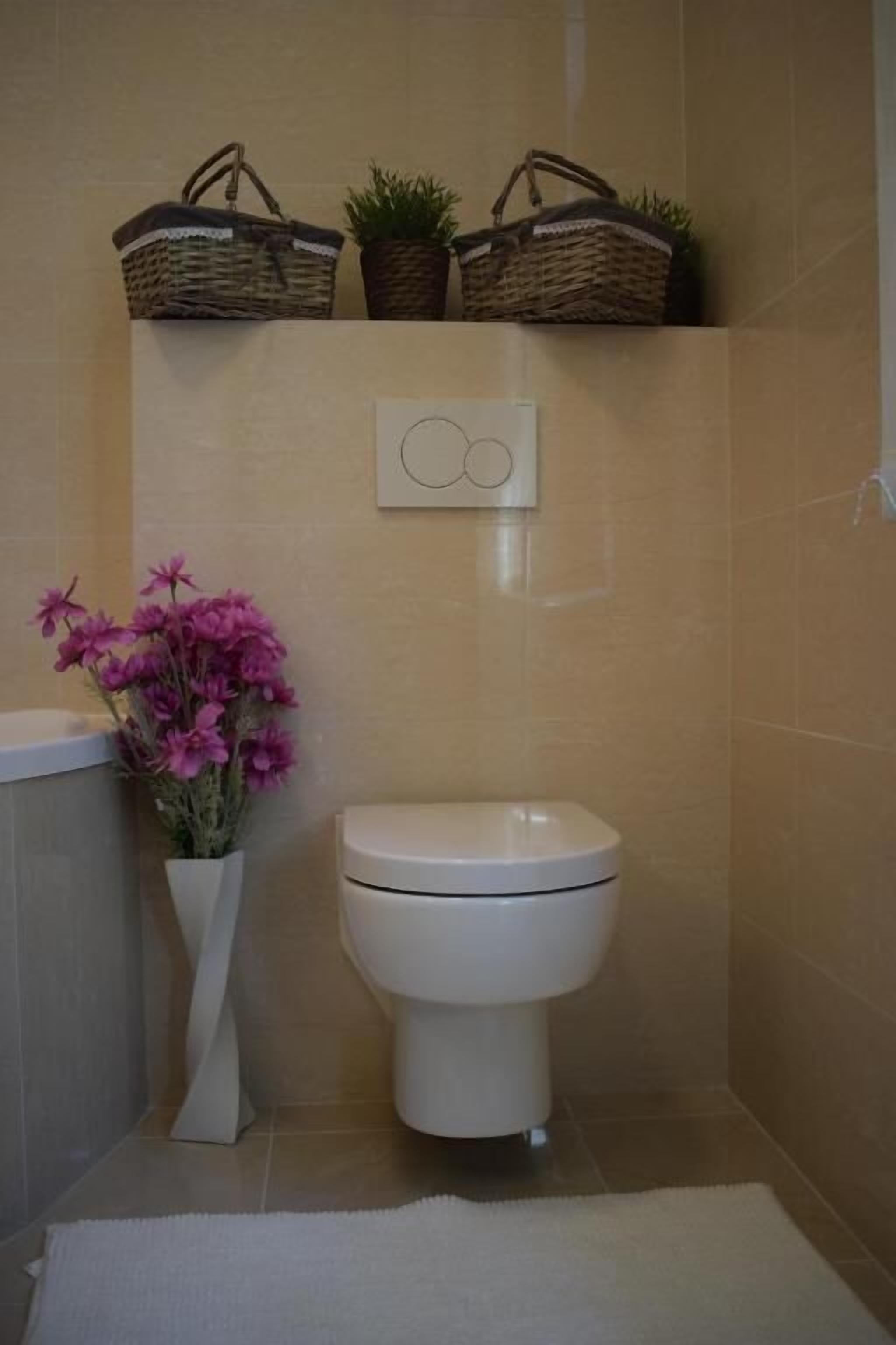 A bathroom with a white toilet, a white vase with pink flowers, and two wicker baskets on the wall.