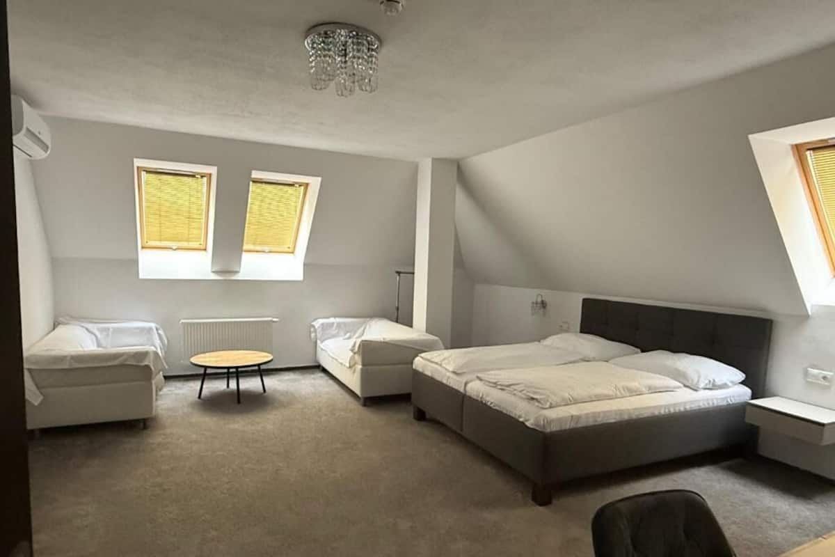 Un dormitorio con una cama grande, dos mesitas de noche, una silla, una pequeña mesa redonda y dos ventanas con persianas.