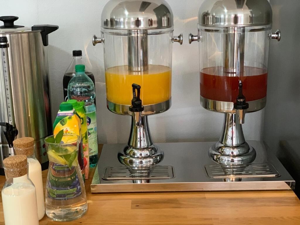Two stainless steel beverage dispensers with transparent containers, a water bottle, and a glass with a straw on a wooden surface.