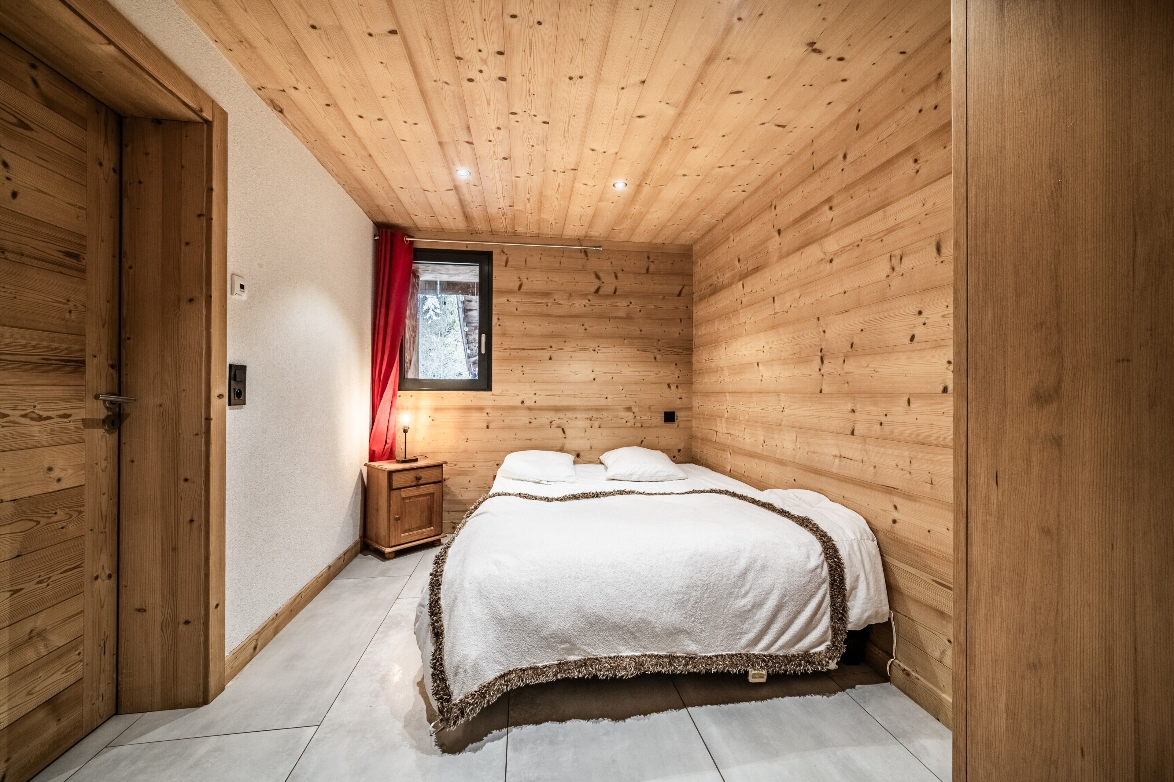 A wooden-paneled bedroom with a bed, a nightstand, a window with a red curtain, and a door.