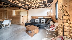 Ein gemütliches Blockhaus-Interieur mit Holzbalkendecke, einer schwarzen Ledersofa, einem Holztisch und einem weißen Metallstuhl.