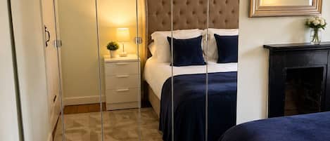 A bedroom with a bed, a nightstand, a lamp, a mirror, and a fireplace.