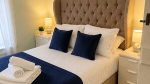 Een netjes opgemaakt bed met een gestoffeerd hoofdbord, twee nachtkastjes met lampen, een decoratieve wandversiering en een kamerbreed tapijt met dessin.