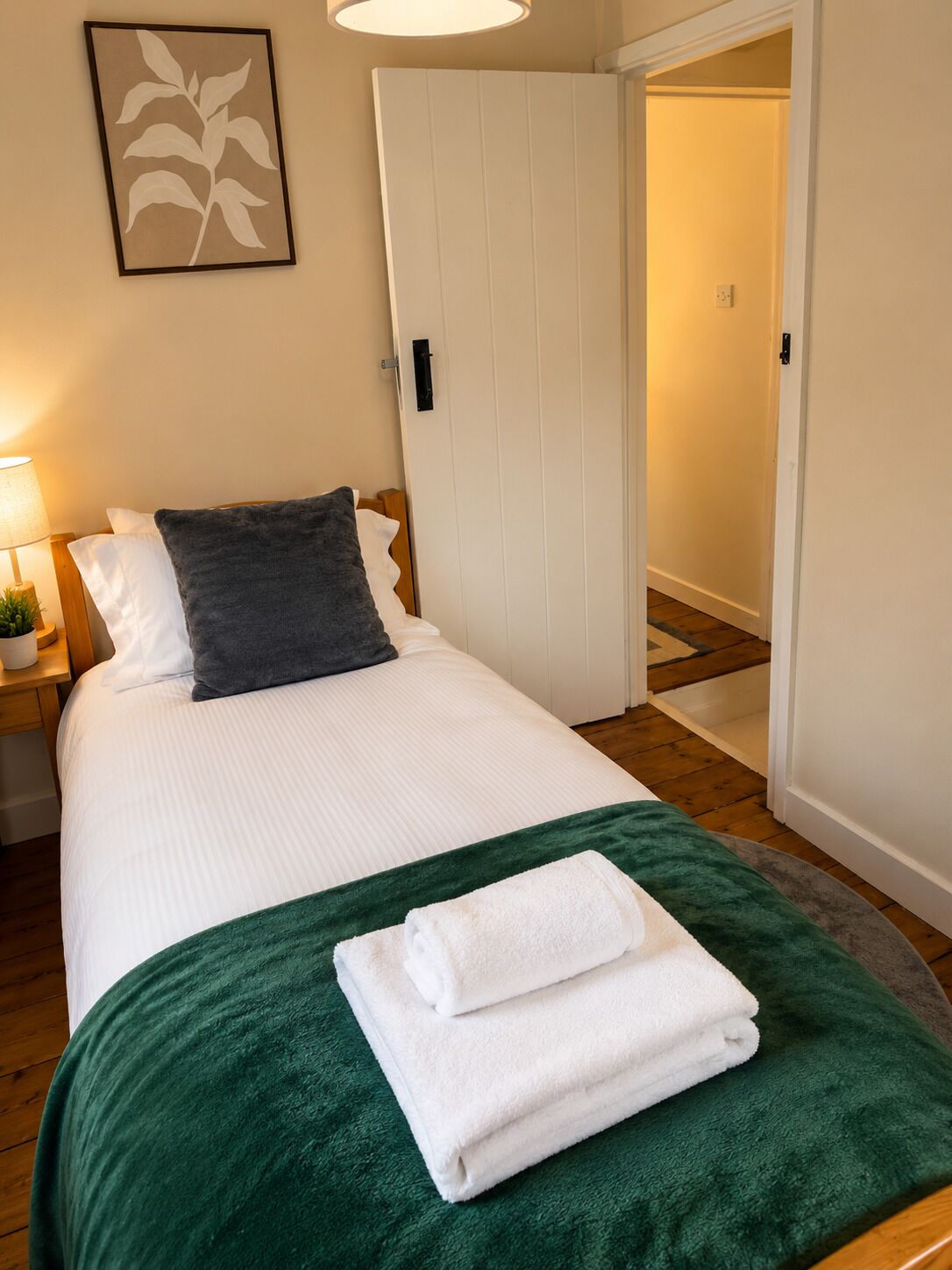A neatly made bed with a green blanket and white towels, a bedside lamp, and a framed picture on the wall.